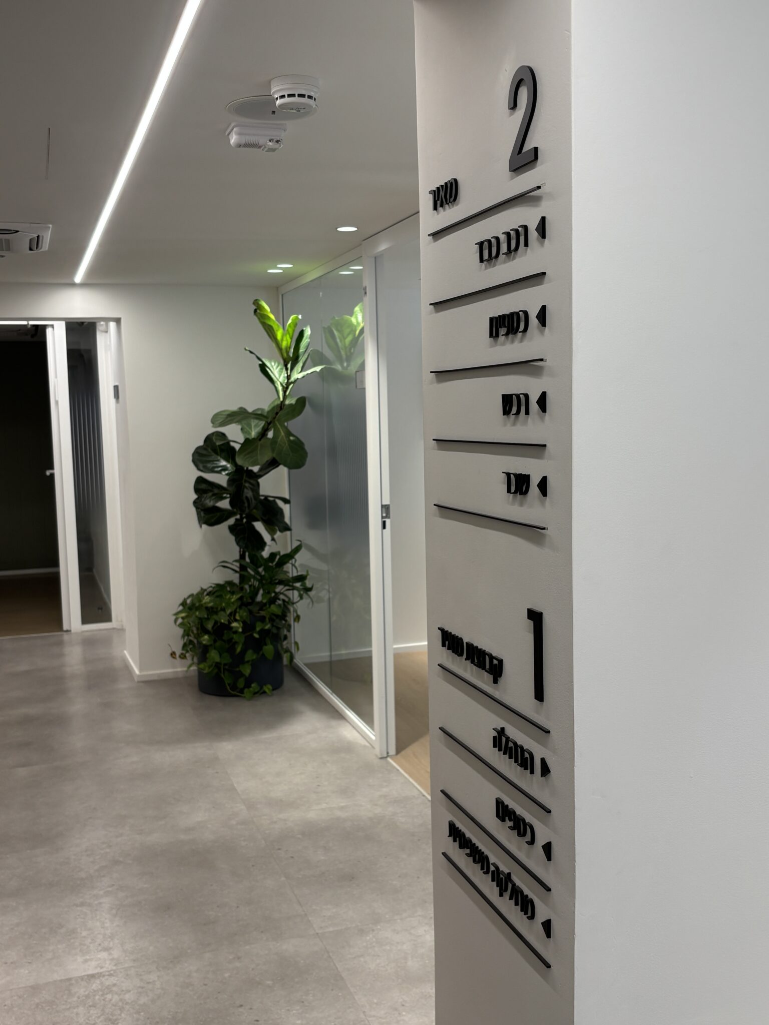 Hebrew floor directory sign beside a corridor with a plant